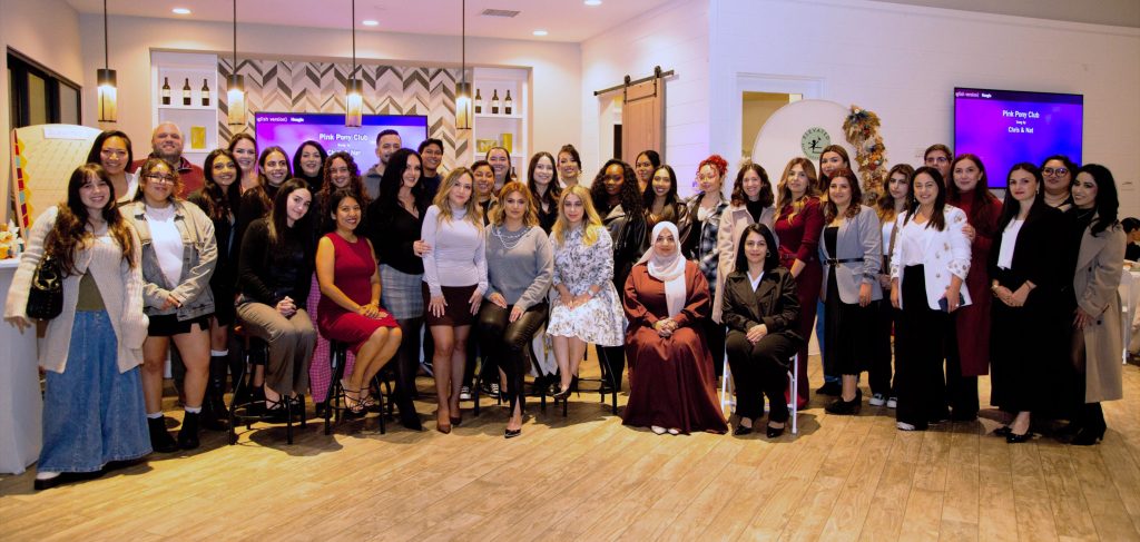 A large group of diverse women and a few men pose together in a well-lit interior space, smiling and dressed in a variety of attire, while a presentation screen in the background displays "Pink Pony Club." The scene conveys a sense of community and celebration.